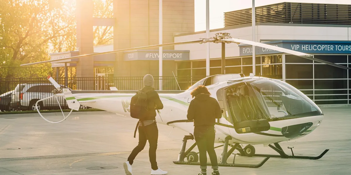 Two people walk toward a small helicopter on a sunlit airport tarmac, evoking themes of flight and travel.