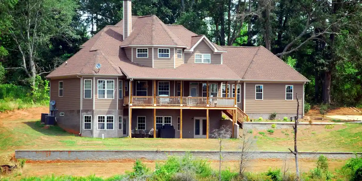 A large brown two-story house with a wooden deck and staircase, set in a yard surrounded by trees