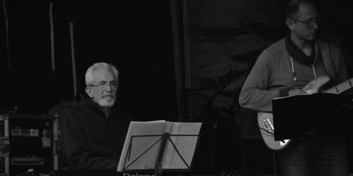 Two musicians performing in a dimly lit setting: a seated pianist with sheet music on a stand, and a standing guitarist with a guitar.