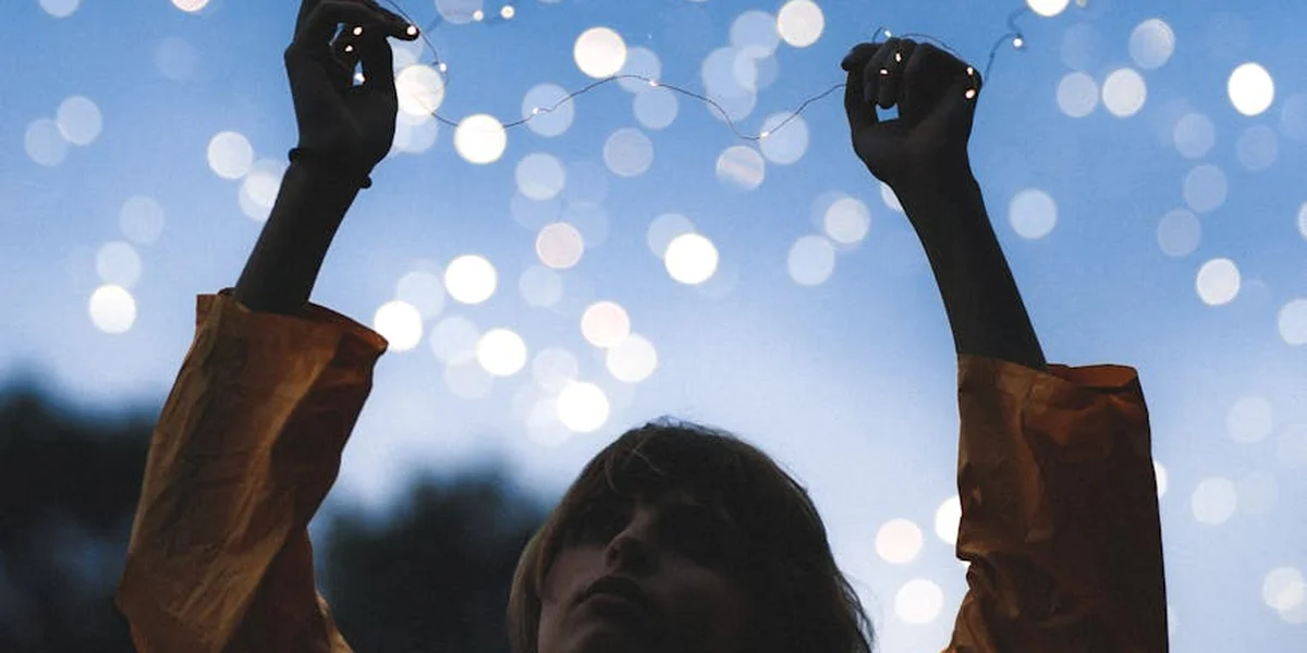 A person wearing a brown jacket raises their arms toward a night sky filled with blurred white lights, creating a dreamlike scene suitable for interpreting dreams.