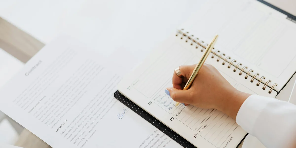 A person writes in a spiral notebook at a desk with a calendar and scattered papers, symbolizing planning for long-term dream empowerment.