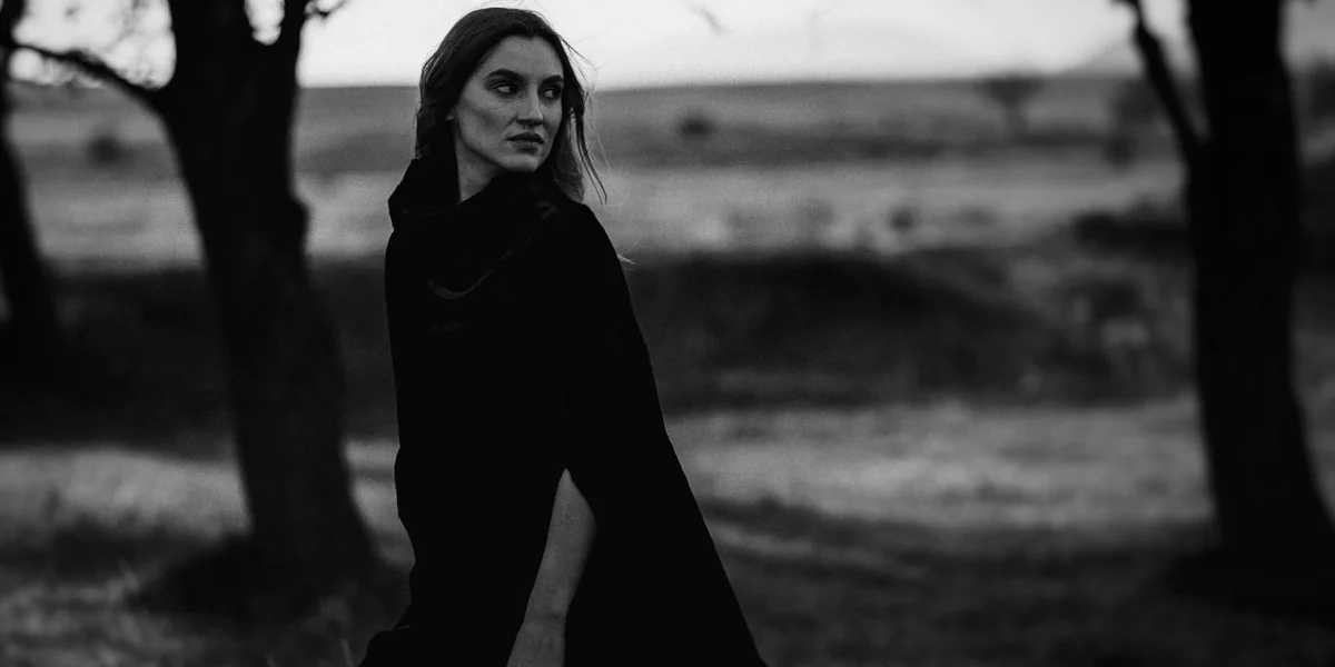 Black-and-white image of a woman walking through a barren field with bare trees, glancing over her shoulder, creating a moody, dreamlike atmosphere.