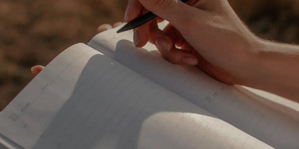 Close-up of a hand holding a pen and writing in an open notebook outdoors