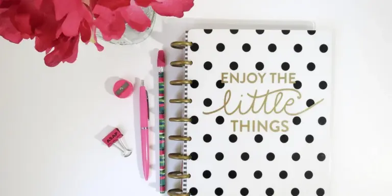 A white desk with a black-polka-dotted spiral notebook featuring gold lettering that reads 'Enjoy the little things', along with pink pens, a pink eraser, and bright pink flowers.