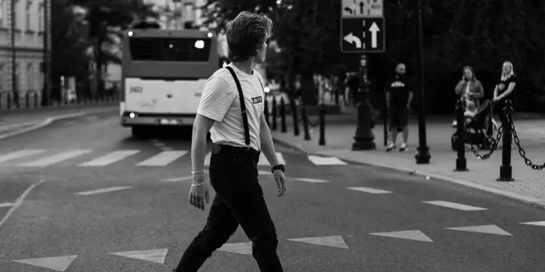 Black-and-white street scene of a person crossing a city crosswalk, wearing a white shirt and suspenders.