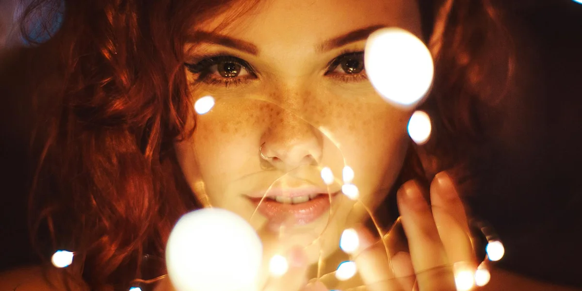 Close-up portrait of a red-haired, freckled woman smiling softly, with soft bokeh lights shimmering around her face.