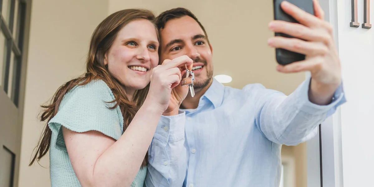 A couple smiles while taking a selfie indoors; the woman holds a small key-like object near the man's face, suggesting the rediscovery of a long-lost item.