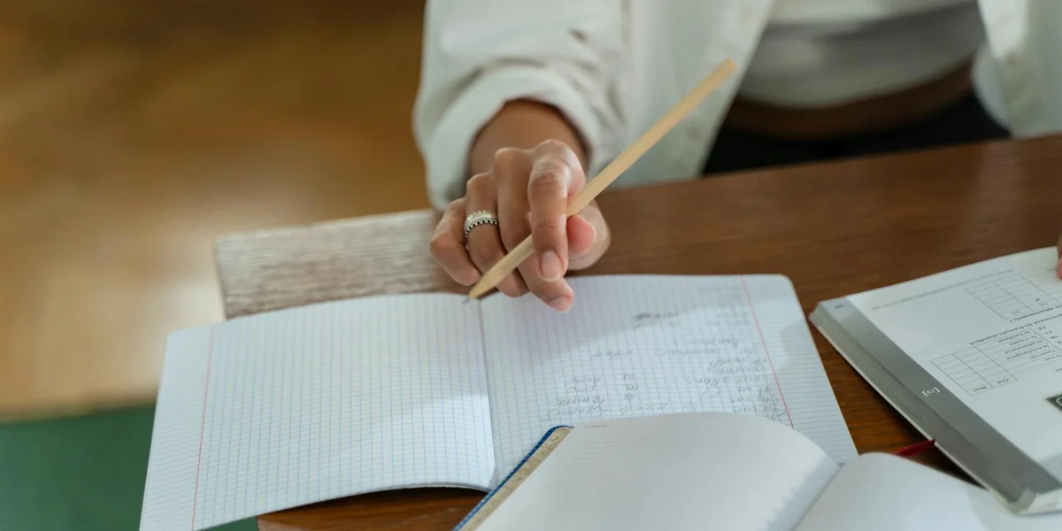 Close-up of a person writing in a notebook on a wooden desk, with papers and a binder nearby.