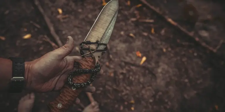 Close-up of a hand gripping a knife with a wrapped handle over a dirt ground.