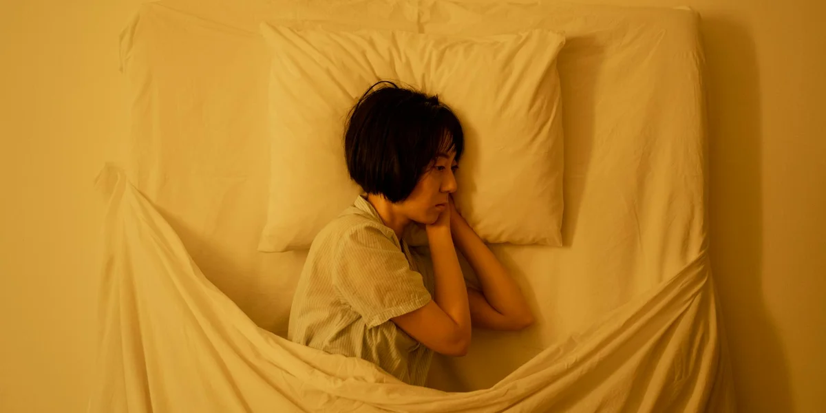 Person lying in bed with hands clasped near the face, appearing distressed in a softly lit room.