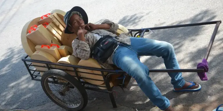 A man wearing jeans and flip-flops reclines on a handcart piled with boxes on a sunny street.