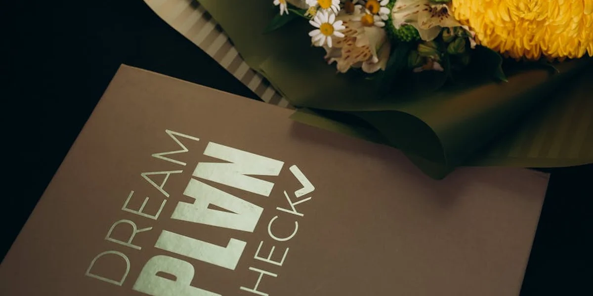 Close-up of a brown card with the words 'DREAM PLAN' in white letters, partially obscured, with a bouquet of white and yellow flowers in the background.