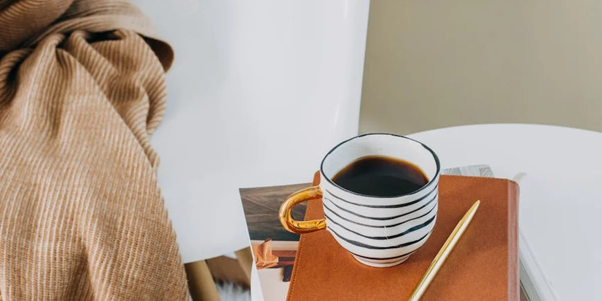 Close-up of a white table featuring a striped coffee mug, a brown notebook, a golden pencil, and a soft beige blanket draped on the left.