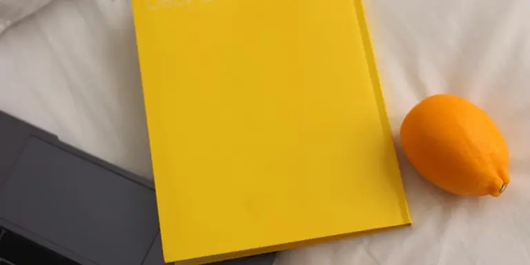 Bright yellow notebook on a white bedsheet with an orange fruit beside it, suggesting morning journaling for dream recall.