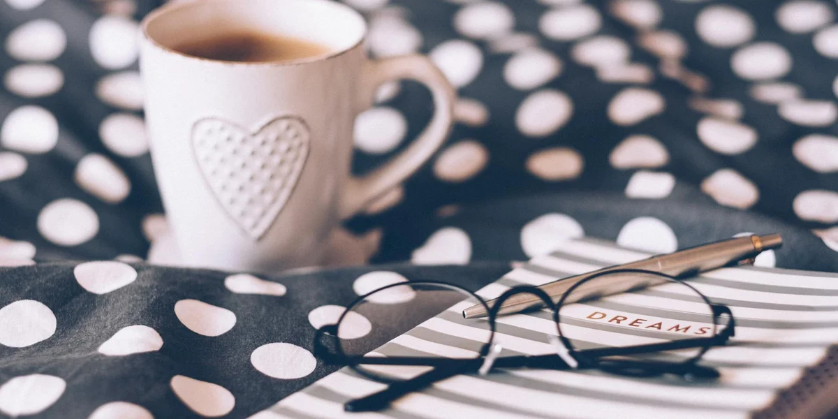 A cozy desk scene with a coffee mug featuring a heart pattern, a pair of reading glasses, and a notebook labeled 'DREAMS' resting on a polka-dotted fabric surface.