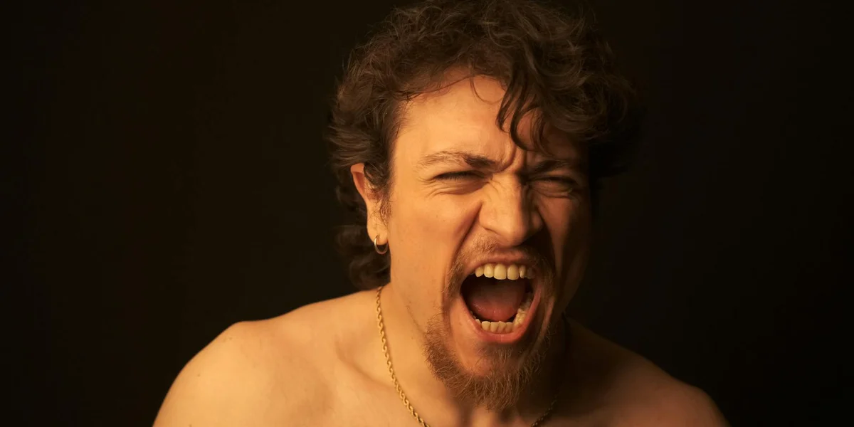 A man with a gold chain screams with his mouth wide open against a dark background, conveying alarm and release.