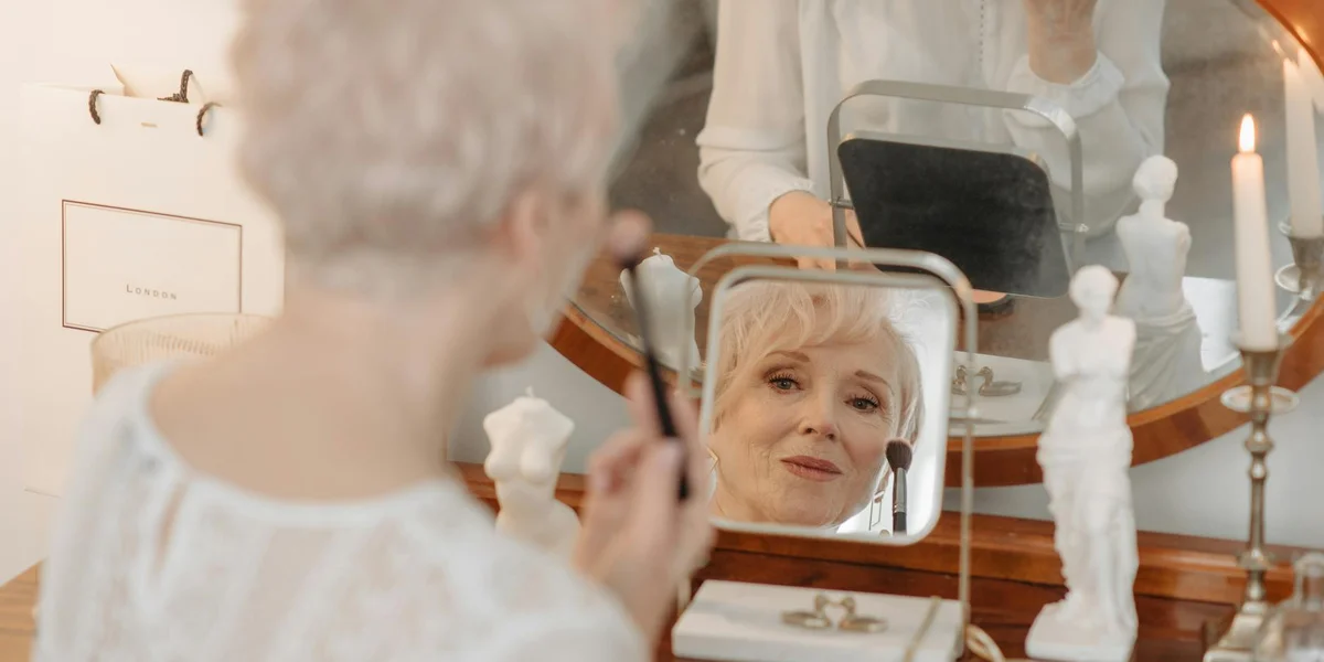 An older woman applying makeup at a vanity, looking into a small handheld mirror.