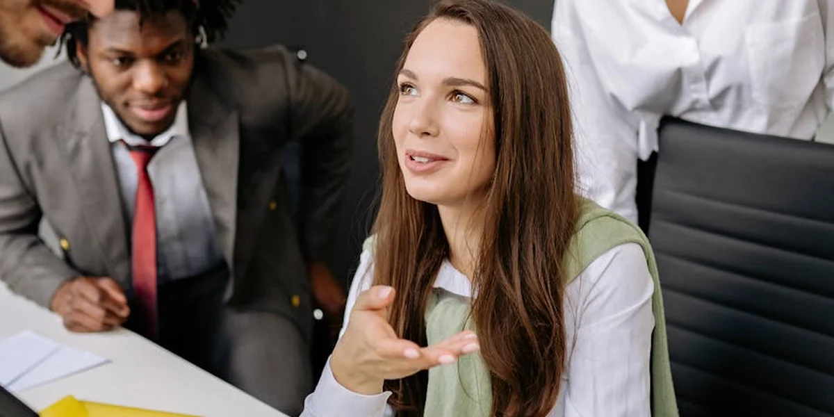 A businesswoman sits at a conference table with a thoughtful expression, while two colleagues lean in, suggesting listening, guidance, and collaborative decision-making.