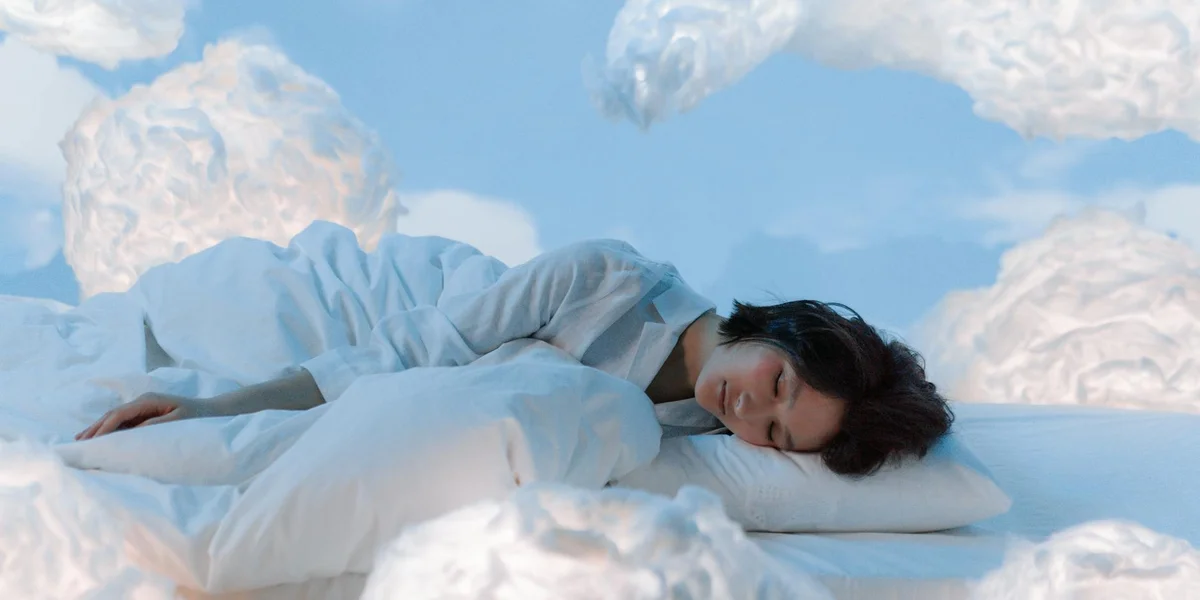 A person sleeping on a white bed surrounded by fluffy clouds in a bright blue sky, creating a surreal dreamscape.