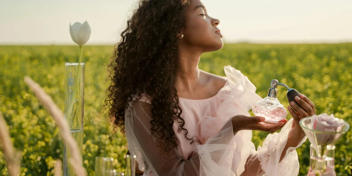 A woman with curly hair stands in a sunlit field of yellow blossoms, holding a perfume bottle and smelling its scent with eyes closed.