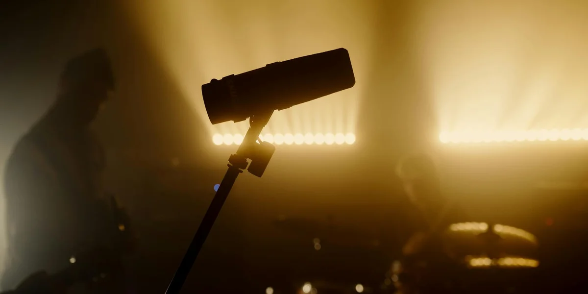 Silhouette of a microphone on a stand against warm backstage lights and hazy atmosphere, with blurred figures in the background.