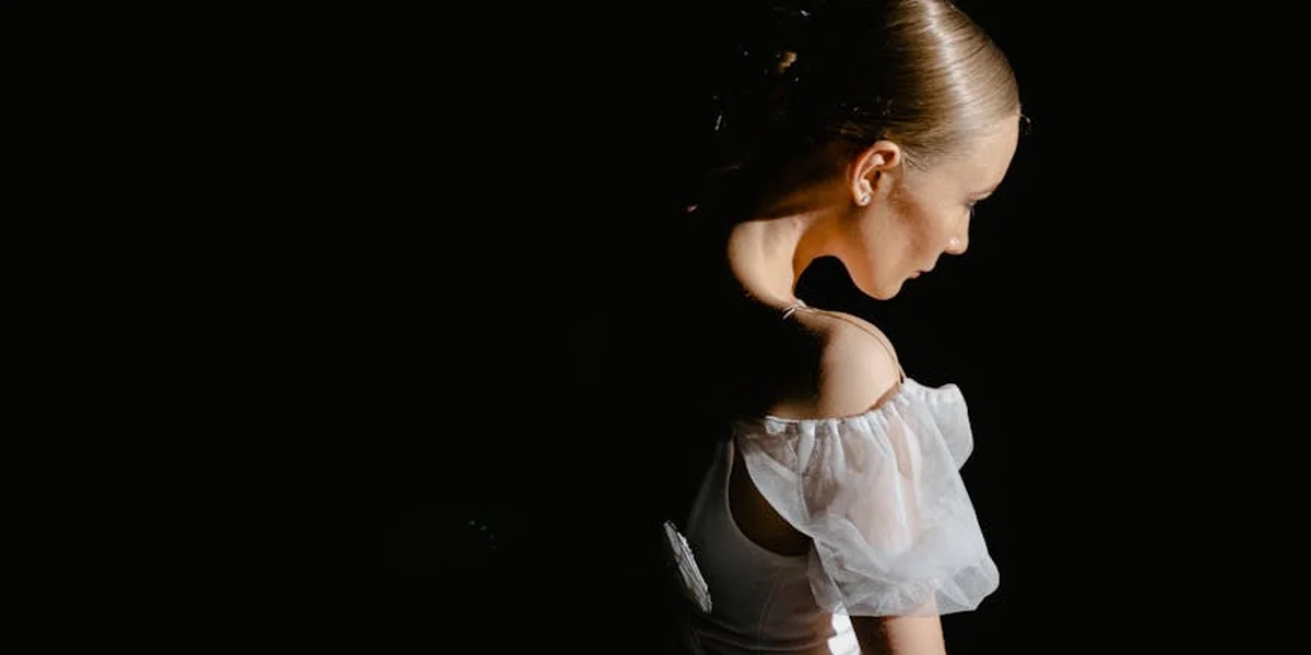 A dancer stands in soft light with a bowed head, wearing a white dress with sheer sleeves, set against a dark stage.