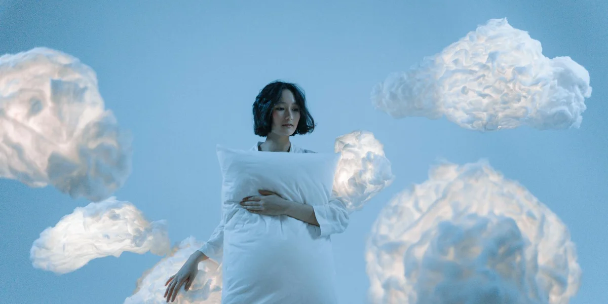 A person cradles a pillow while fluffy clouds drift in a clear blue sky.