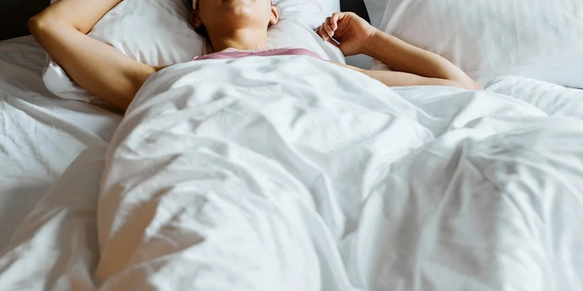 Person lying in bed under white sheets, arms resting above the head in a calm bedroom setting
