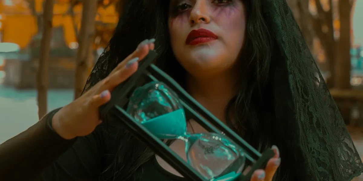Portrait of a person with dark hair and red lipstick holding an hourglass with turquoise sand, set against a moody, out-of-focus background.
