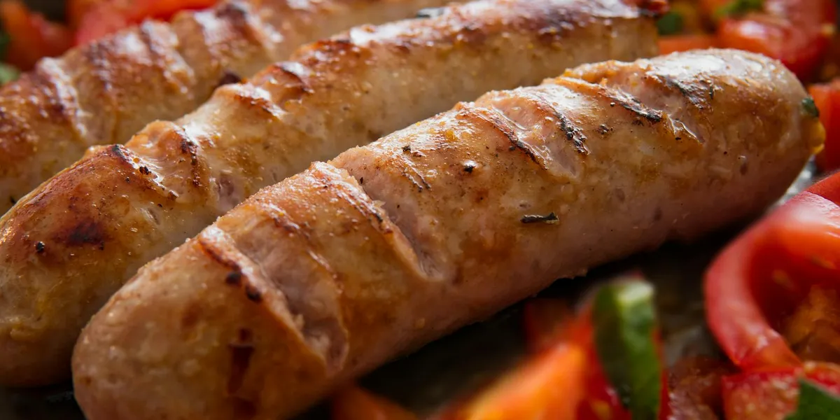 Close-up of grilled sausages with herbs and vegetables in the background, including red tomato slices.