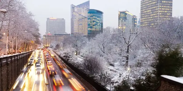 Snow-covered city street with vehicles stuck in traffic, flanked by leafless trees and tall modern buildings in the background.