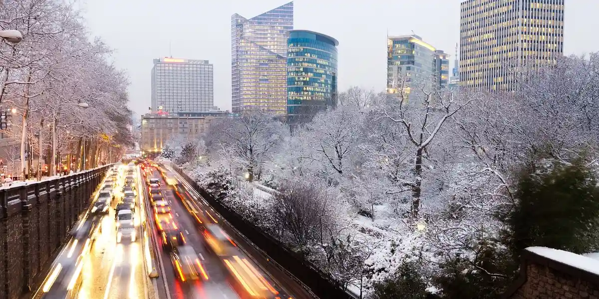 Snow-covered city street with vehicles stuck in traffic, flanked by leafless trees and tall modern buildings in the background.