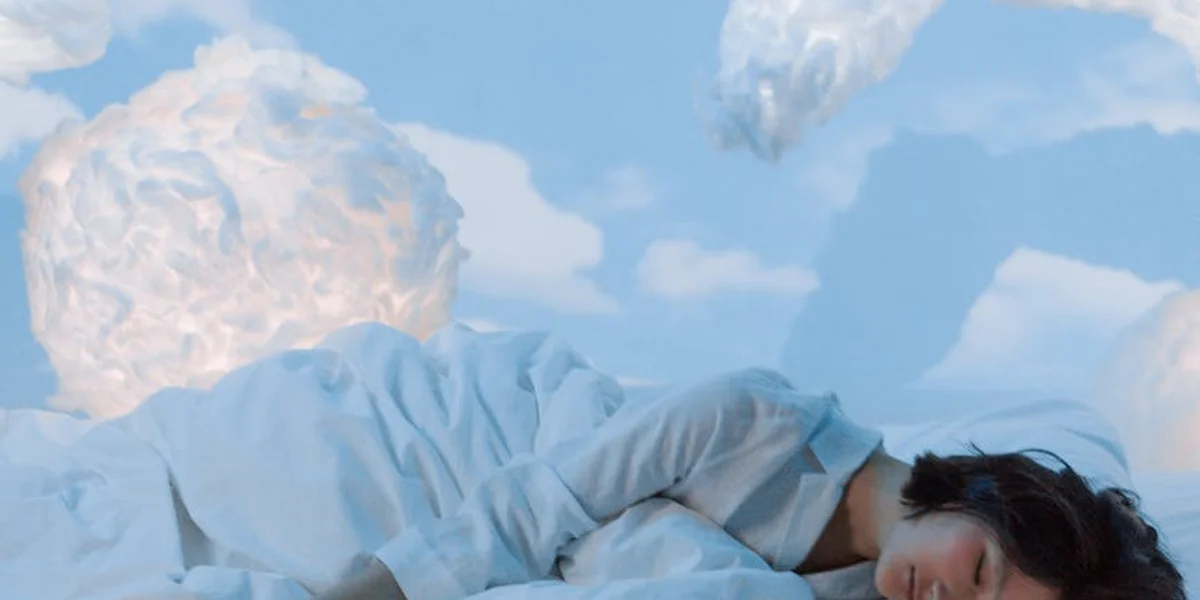 A person in a light blue shirt lying on a bed that blends into a sky filled with clouds, symbolizing dreaming and decoding unfamiliar imagery.