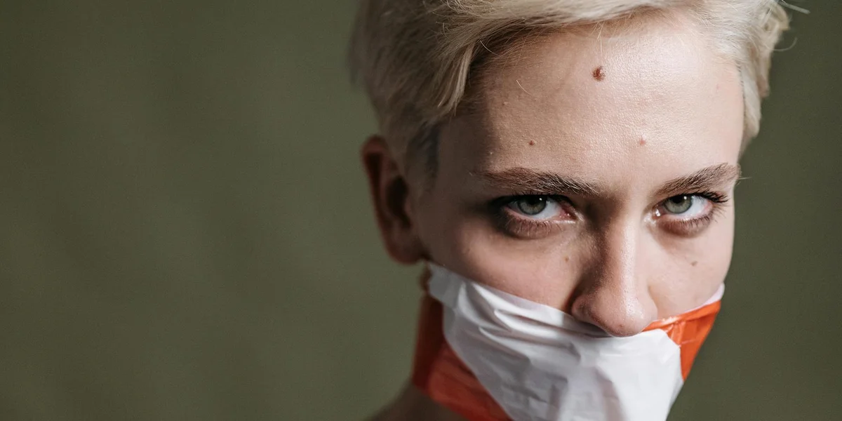 Close-up of a person with a mask covering the mouth and intense eyes, short blond hair, conveying silence and constraint.