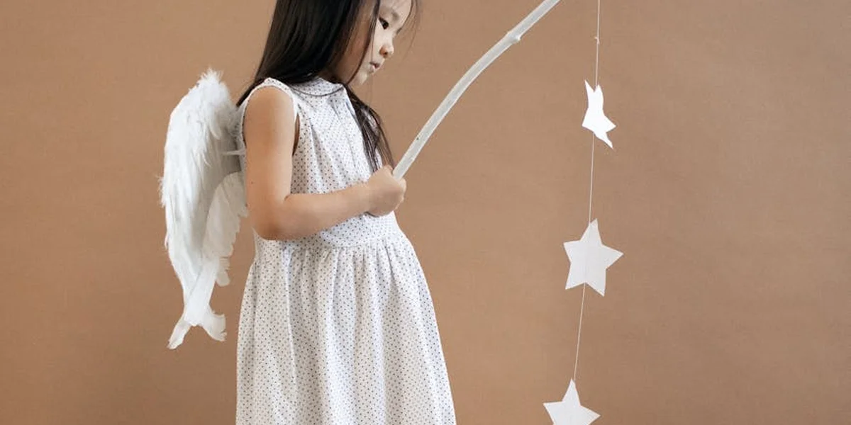 A young girl with white angel wings wearing a white polka-dot dress stands against a warm brown background, holding a curved white object resembling a bow, with a string of hanging stars beside her.