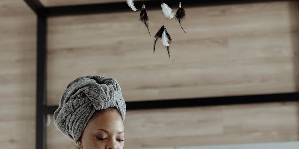 Portrait of a person wearing a gray headwrap, head bowed, with a wooden paneled background and hanging feathers above, evoking a loom-like, contemplative atmosphere.