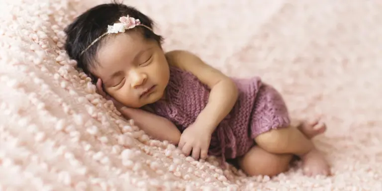 Sleeping newborn wearing a purple knitted outfit and a delicate flower headband, resting on a soft pink blanket.