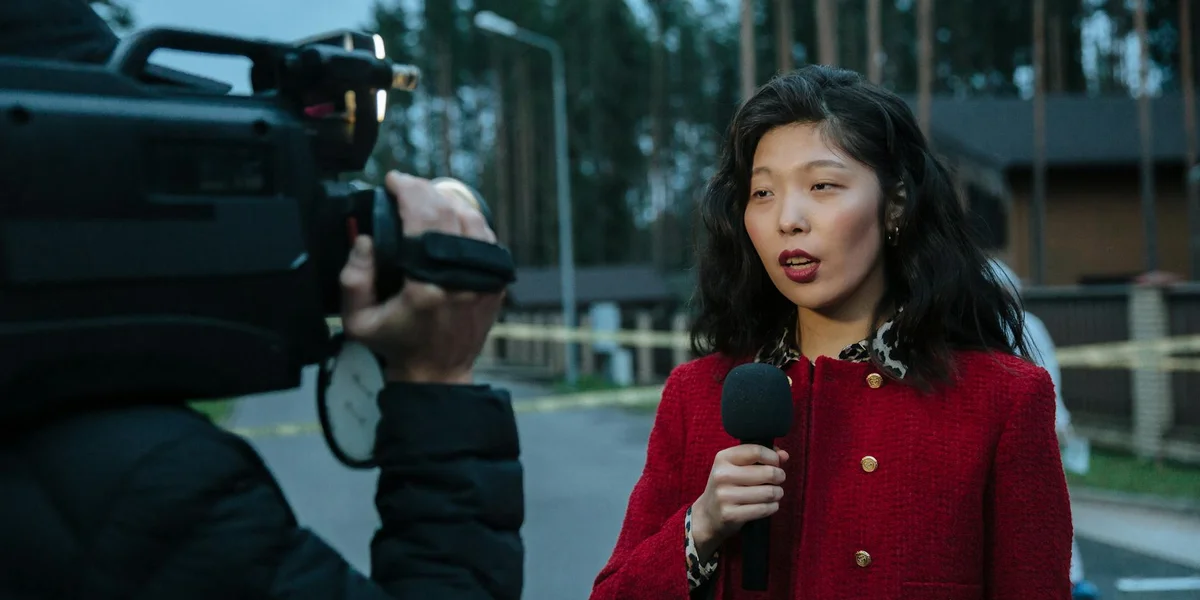 A woman with shoulder-length dark hair wearing a red jacket speaks into a handheld microphone, while a camera operator films her. They stand near a cordoned-off crime scene with yellow tape in the background.