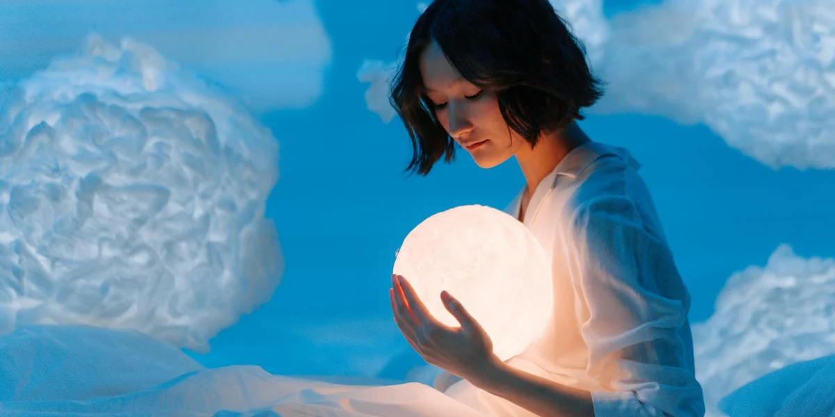 A person holding a glowing orb against a bright blue sky with fluffy clouds, symbolizing the unconscious shaping dreams.