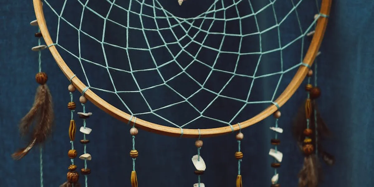 Close-up of a dreamcatcher with a woven web and hanging feathers against a blue background.
