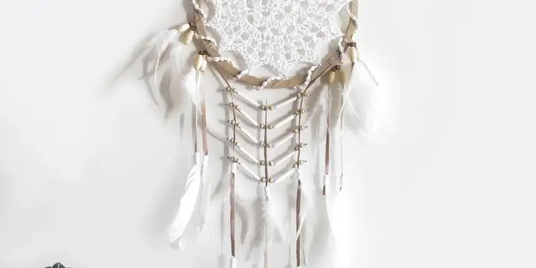 Close-up of a white lace dreamcatcher necklace with feathers and beads on a pale background.