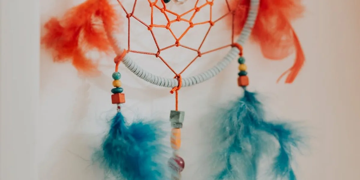 Close-up of a dreamcatcher with an orange web, a pale gray hoop, turquoise beads, and blue feathers hanging from the bottom.