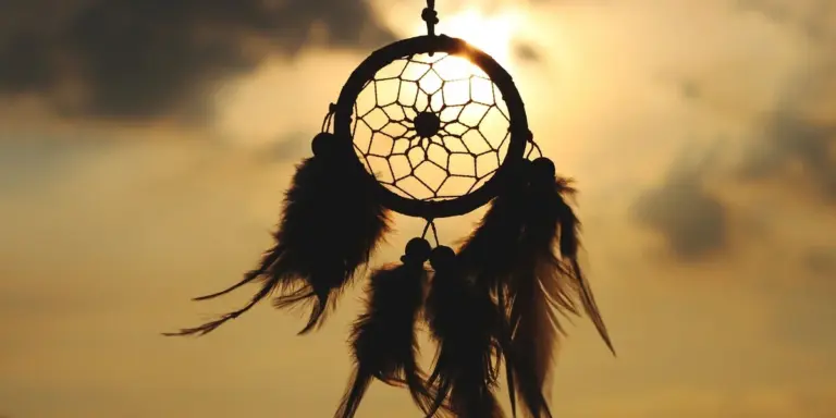 Silhouette of a dreamcatcher hanging against a warm sunset sky, with feathered strands gently swaying.