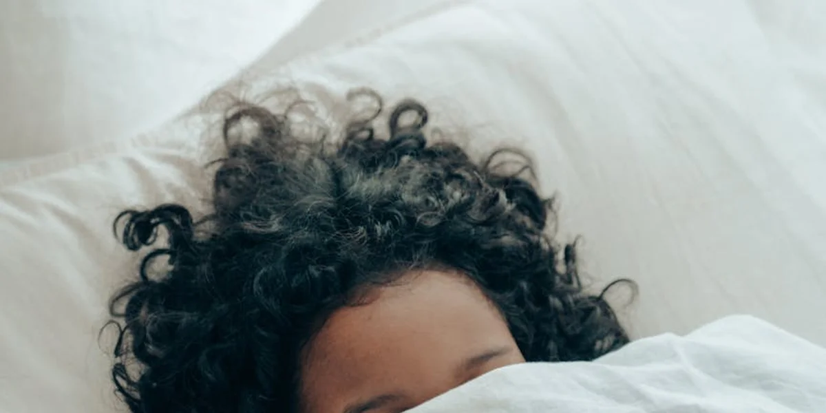 Close-up of a person with curly hair sleeping in bed under white sheets.