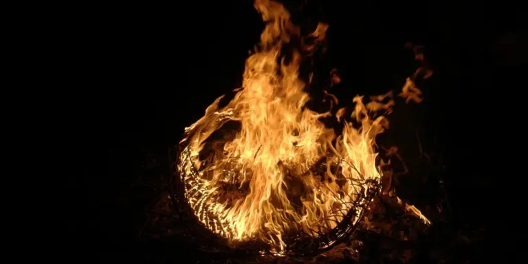 A bright campfire burning against a dark night, with orange and yellow flames curling around charred wood.