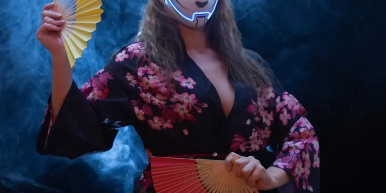 A person wearing a decorative mask and a floral kimono holds a folding fan against a smoky blue background.