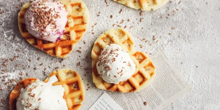 Several heart-shaped waffles topped with scoops of ice cream and chocolate shavings on a light textured surface.