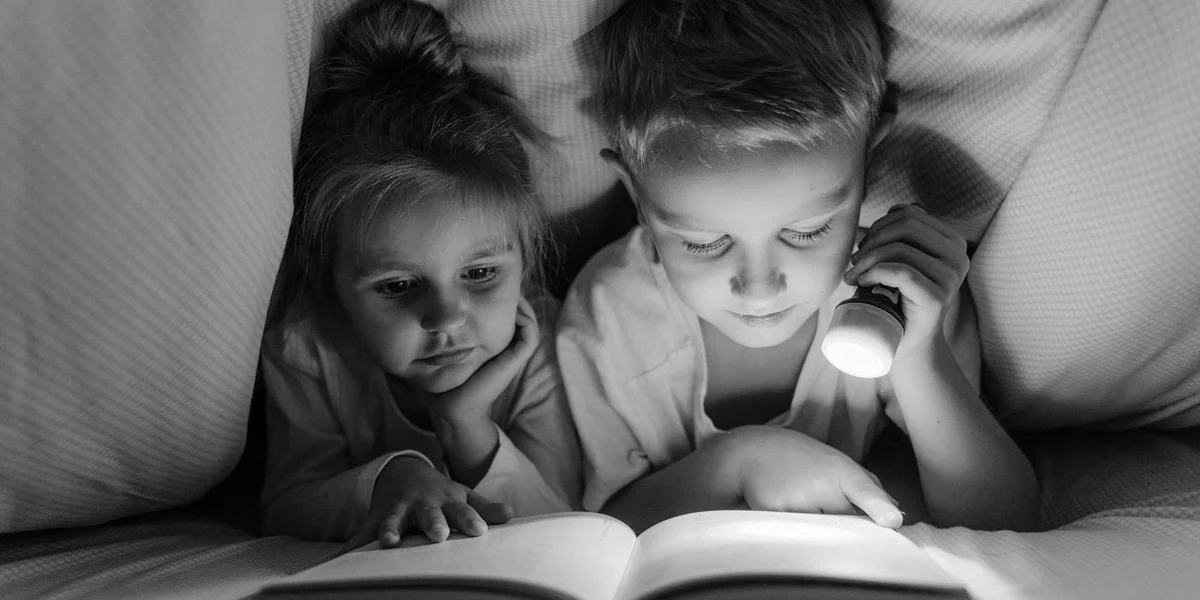 Two children snuggled under a blanket, one shining a flashlight on a book as they read together in black and white.