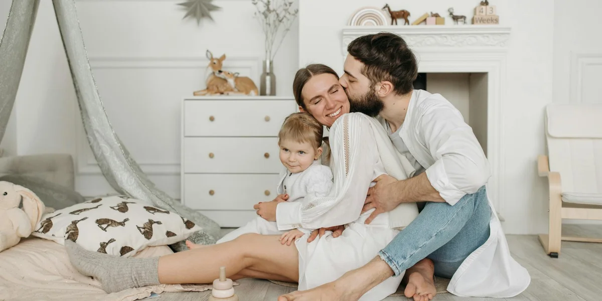 A couple hugging their young child in a bright, cozy bedroom, smiling warmly.