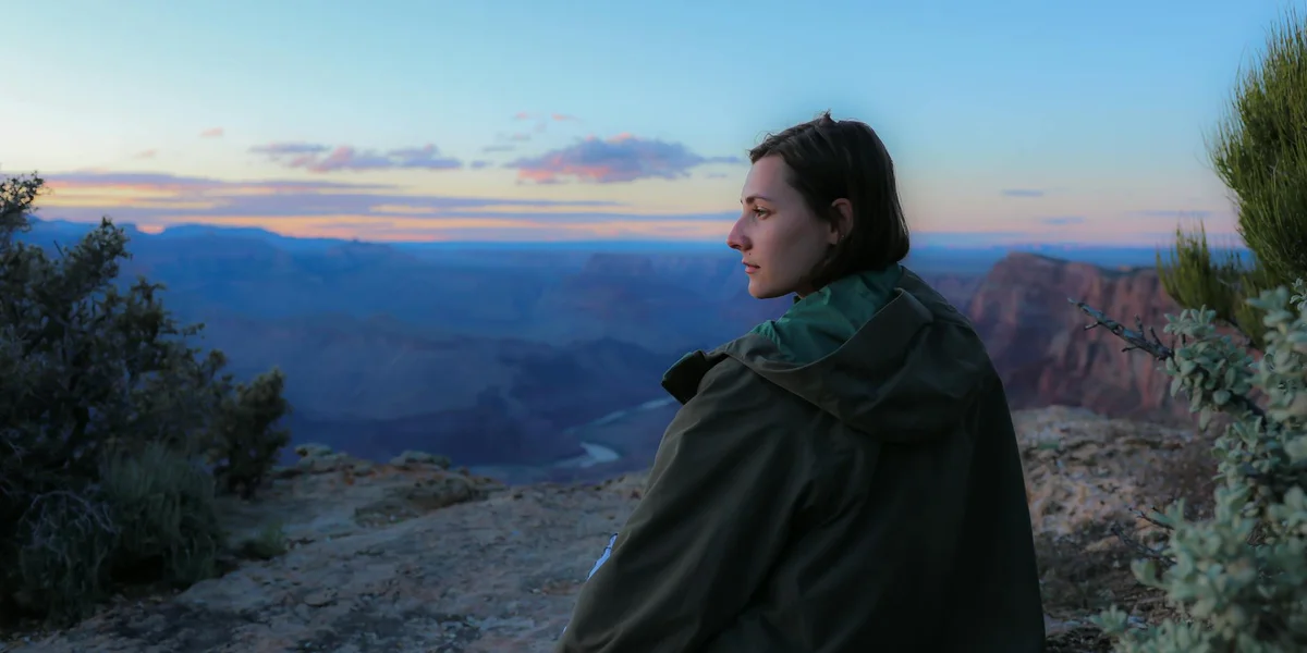 Person standing on a cliff overlooking a canyon at sunset, wearing a dark coat and looking toward the horizon.